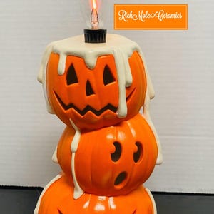 Könnte beinhalten: Gestapelte Halloween-Kürbisdekoration mit Glühbirne. Die orangefarbenen Keramikkürbisse haben geschnitzte Gesichter und tropfen mit weißem Wachs. Der Text "Rich Mole Ceramics" befindet sich in einem orangefarbenen Rechteck.