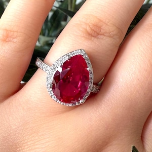 Puede incluir: Un anillo de plata con una gran piedra preciosa roja en forma de pera, rodeada de piedras más pequeñas y transparentes. El anillo está en una mano con uñas cuidadas, con follaje verde en el fondo.