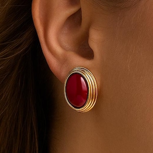Może przedstawiać: Złote kolczyki owalne z żywym, czerwonym kamieniem centralnym. Kolczyki mają warstwowy wzór wokół czerwonego kamienia. Obraz przedstawia zbliżenie kolczyków na uchu.