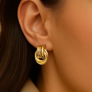May include: Gold-colored stud earrings with a looped design. The earrings are made of a shiny, metallic material and are worn on the earlobe. The earrings have a classic, elegant style.