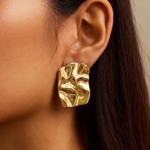 May include: Gold-toned square earrings with a textured, organic design. The earrings have a molten, rippled appearance, reflecting light. These statement earrings are a bold accessory.