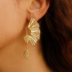 Puede incluir: Pendientes dorados con una parte superior en forma de abanico y tres colgantes con forma de hoja. Los pendientes se llevan en una oreja, con el diseño que capta la luz. Los pendientes son una pieza llamativa.