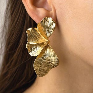 May include: Gold-toned statement earrings with a textured, floral design. The earrings feature three overlapping petals, creating a bold and elegant look. These earrings are a stylish accessory for any occasion.