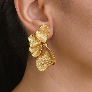 May include: Gold-toned statement earrings featuring a textured, leaf-inspired design. The earrings have a three-dimensional, openwork structure, with multiple overlapping sections. The earrings are worn on the ear, and the image shows a close-up of the ear and the jewellery.