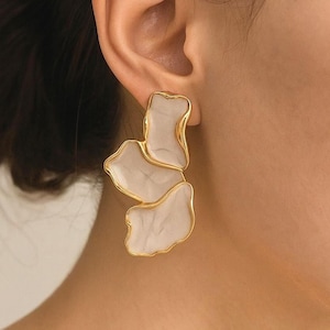May include: Gold-toned statement earrings with a layered, abstract design. Each earring features three irregular, petal-like shapes in a marbled white, outlined with a gold border. The earrings are shown on an ear.
