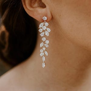 May include: Elegant, long dangle earrings with a floral design. The earrings feature multiple clear, sparkling crystals set in a gold-toned metal. The design includes round and marquise-shaped stones, creating a cascading, leaf-like pattern. A sophisticated accessory.