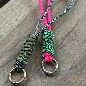 May include: Two handmade lanyards with metal rings. One is olive green with a gold ring, the other is green and blue with a silver ring. Both have a loop of patterned cord and a bright pink cord.