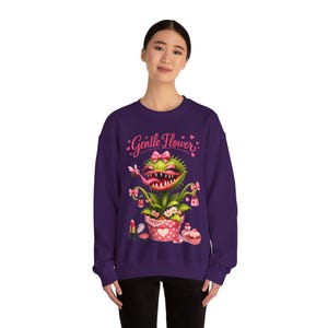 May include: A purple sweatshirt featuring a cartoon plant with a large mouth, pink bow, and lipstick. The text "Gentle Flower" is above the plant. The plant is in a pink polka-dot pot with makeup.