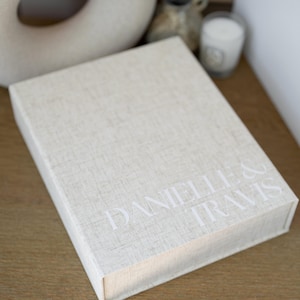 May include: A rectangular keepsake box covered in a light beige, textured linen-like fabric. The top surface features the white printed text DANIELLE & TRAVIS in a clean, sans-serif font. The box has a hinged lid and a structured, boxy shape.
