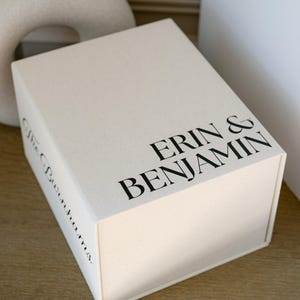 Puede incluir: Una caja rectangular de color blanco hueso con cierre magnético que presenta el texto impreso en negro ERIN & BENJAMIN en la tapa superior y el texto en cursiva The Burnhams impreso en negro en el panel lateral de la caja.