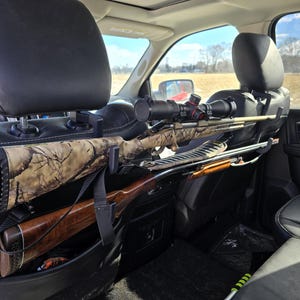 May include: A black car seat gun rack holding two rifles. One rifle has a camouflage pattern, and the other has a wooden stock. A scope is mounted on the top rifle. The rack is attached to the headrest of the car seat.
