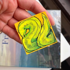 May include: A small, square sticker with a vibrant yellow background featuring a detailed illustration of a green moray eel. The eel is depicted in a stylized, artistic manner with dark green outlines and shading.