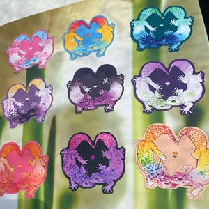 May include: A collection of colorful gecko stickers. Each sticker features two geckos in a heart shape, with various color schemes including pink, blue, purple, and rainbow. Some stickers have a small rainbow heart symbol.