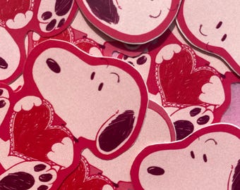 May include: A collection of Snoopy stickers with red heart designs. The stickers feature Snoopy's face and paw prints, with a red heart in the center. The stickers have a pink background and a red outline.