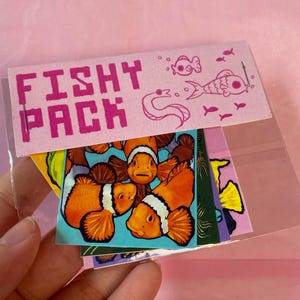 May include: A clear plastic sleeve holding a pack of colorful stickers. The pink header reads "Fishy Pack" with fish illustrations. The stickers feature clownfish, and other aquatic designs.