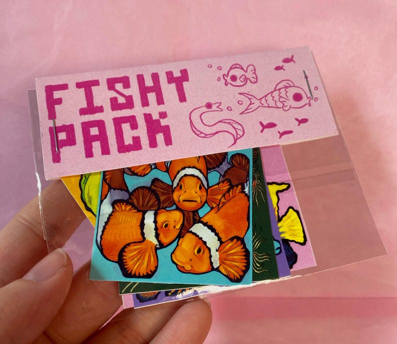 May include: A clear plastic sleeve holding a pack of colorful stickers. The pink header reads "Fishy Pack" with fish illustrations. The stickers feature clownfish, and other aquatic designs.
