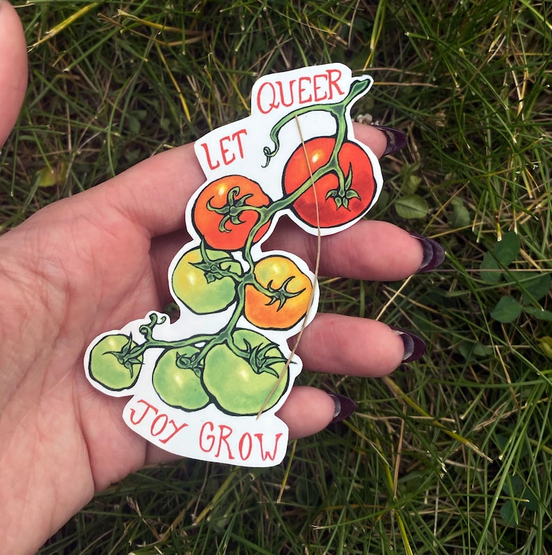 May include: A sticker depicting a cluster of tomatoes, ranging in colour from green to red. The words "LET QUEER JOY GROW" are printed on the sticker in a playful font. The sticker is held against a background of green grass.