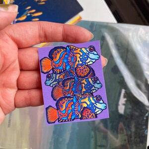 May include: A sticker featuring a stack of four colorful mandarin fish illustrations. The fish have orange, blue, and yellow patterns on a purple background. The sticker is held by a hand.