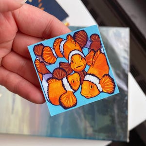 May include: A square sticker featuring a group of cartoon clownfish. The fish are orange with white stripes and black-edged fins, set against a light blue background. The sticker is held by a hand.
