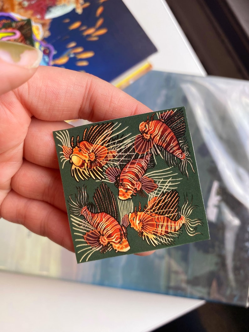 May include: A square sticker featuring five orange and white lionfish on a dark green background. The lionfish have distinctive fan-like fins and striped bodies. The sticker is held in a hand.