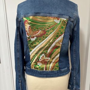 Op de afbeelding: Blauwe spijkerjas met een decoratief paneel op de rug. Het paneel heeft een gouden, groene en rode vlinderdesign. De jas heeft een klassieke kraag, knoopsluitingen en lange mouwen.