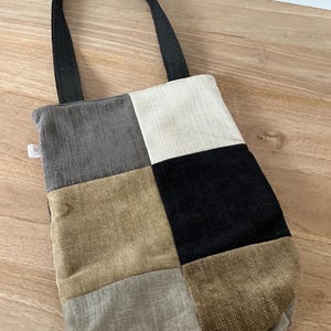 Op de afbeelding: Een patchwork tote bag met een zwarte handgreep. De tas is gemaakt van stoffen vierkanten in tinten grijs, beige en zwart. De tas is ontworpen voor het dragen van spullen en heeft een eenvoudig, functioneel ontwerp.