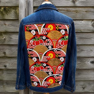 Op de afbeelding: Een donkerblauw spijkerjack met een groot rechthoekig achterpaneel met een levendig, herhalend patroon van gouden, rode, witte en zwarte waaiervormen, bloemmotieven en geometrische ontwerpen die in de stof zijn verwerkt.