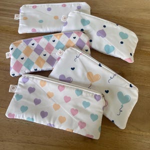Op de afbeelding: Vijf rechthoekige etuis van stof met ritssluiting. De ontwerpen bevatten witte achtergronden met pastelkleurige hartjespatronen, een ruitpatroon in zachte kleuren en een patroon met hartvormige ballonnen met kleine donkerblauwe hartjes.