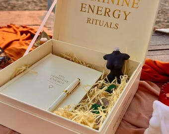 May include: A cream-colored box with the words "THE FEMININE ENERGY RITUALS" in gold lettering. Inside, a journal, pen, and a black torso-shaped candle rest on straw. The journal and pen are white with gold accents. The box is open, revealing the contents.