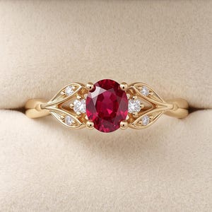 Pode incluir: Um anel de ouro com um rubi vermelho profundo de corte oval ao centro. A banda apresenta motivos metálicos em forma de folha em ambos os lados, cada um acentuado com dois pequenos diamantes redondos transparentes, criando um visual simétrico.
