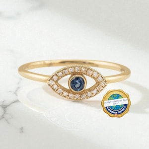 May include: A gold ring featuring an eye-shaped centerpiece. The center of the eye is a round blue stone, surrounded by a halo of small clear stones. A small circular gold badge with a blue globe graphic and the text DELIVERED DUTY PAID DDP is present.