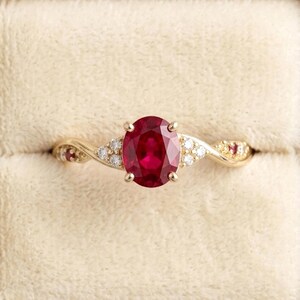 May include: A gold ring featuring a central oval-cut red ruby gemstone, flanked by small round white diamonds and additional tiny red rubies set along the twisted band, presented in a square, cream-colored velvet ring box with a plush interior.