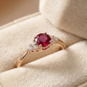 May include: A gold ring featuring a large, round, red gemstone as the central focus. The ring is adorned with smaller, clear gemstones flanking the main stone. Presented in a beige ring box.