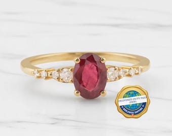 May include: A gold ring featuring a central oval-cut red ruby gemstone flanked by three small round-cut white diamonds on each side of the band. The ring is accompanied by a circular gold seal with a globe graphic and the text DELIVERED DUTY PAID DDP.