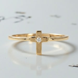 Peut inclure: Une fine bague en or dotée d'une petite pièce centrale en forme de croix. Le centre de la croix est orné d'une unique pierre précieuse claire, taillée en rond, qui scintille. L'anneau est lisse, poli et conserve une largeur fine et uniforme.