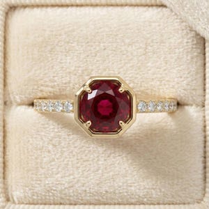 May include: A gold ring featuring a central octagonal-cut deep red ruby held by eight prongs. The band is crafted from yellow gold and is set with a series of small, round, clear white diamonds along both sides of the central gemstone.