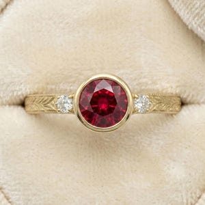 May include: A yellow gold ring featuring a round, deep red ruby set in a bezel setting at the centre. The band is engraved with a leaf-like pattern and includes a small, round, clear white diamond on each side of the central ruby gemstone.