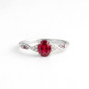 May include: A silver-toned ring featuring a central oval-cut red gemstone held by four prongs. The band has a twisted design accented with small, clear, round-cut stones and two tiny red stones embedded on each side of the central setting.