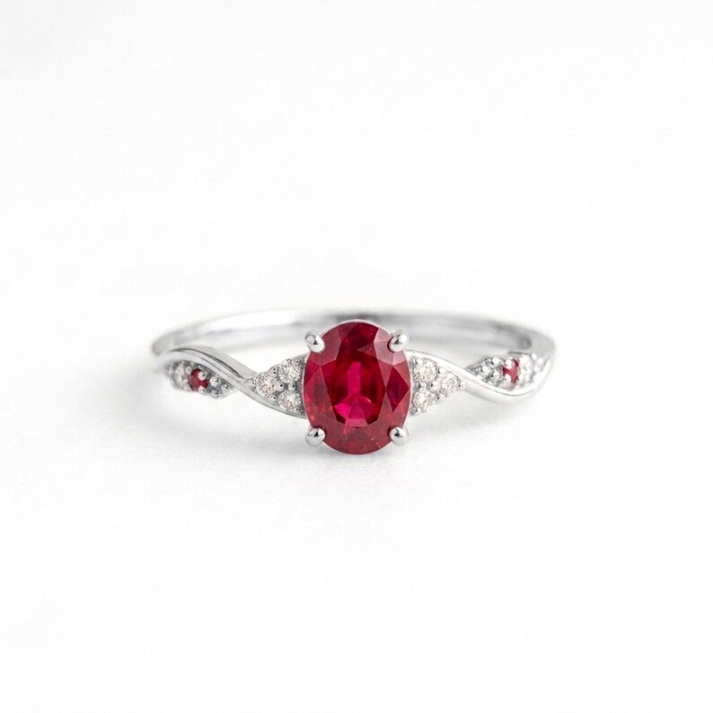 May include: A silver-toned ring featuring a central oval-cut red gemstone held by four prongs. The band has a twisted design accented with small, clear, round-cut stones and two tiny red stones embedded on each side of the central setting.