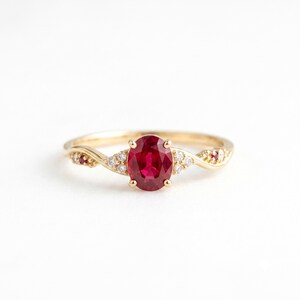 May include: A gold ring featuring a central oval-cut red ruby held by four prongs. The band is accented with small, clear round-cut diamonds and two smaller red rubies set into the gold shank on either side of the primary gemstone.