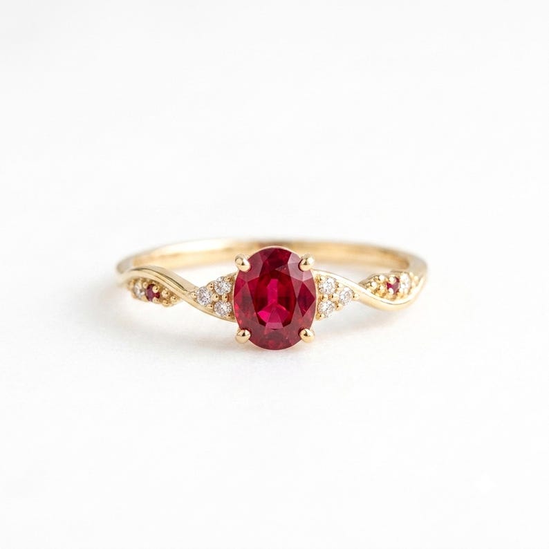May include: A gold ring featuring a central oval-cut red ruby held by four prongs. The band is accented with small, clear round-cut diamonds and two smaller red rubies set into the gold shank on either side of the primary gemstone.