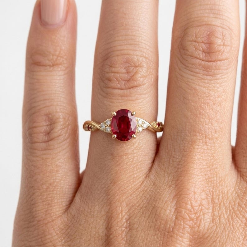 May include: A gold ring featuring a central oval-cut red ruby gemstone held by four prongs. The band has a twisted design and is accented with small, round, clear white diamonds set along the shoulders on both sides of the centre stone.