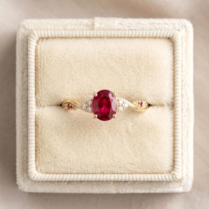 May include: A gold ring featuring a central oval-cut red ruby gemstone, flanked by small round white diamonds and additional tiny red rubies set along the twisted band, presented in a square, cream-colored velvet ring box.