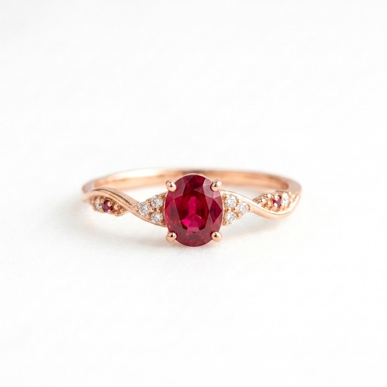 May include: A rose gold ring featuring a central oval-cut red ruby held by four prongs. The band has a twisted design accented with small round white diamonds and smaller red rubies embedded along the sides of the metal shank.