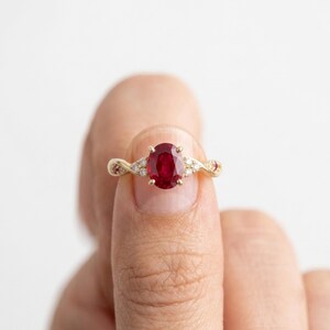 May include: A gold ring featuring a central oval-cut red ruby gemstone held in a four-prong setting. The band has a twisted design and is accented with small, clear round-cut diamonds and two smaller red rubies on either side of the centre stone.