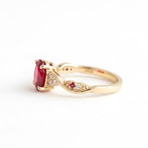 May include: A gold ring featuring a large oval-cut red gemstone as the centrepiece. The band has a twisted design accented with smaller round-cut white diamonds and tiny red gemstones embedded along the sides of the gold metal shank.
