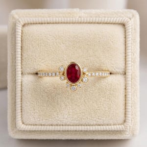 Könnte beinhalten: Ein Goldring mit einem zentralen, oval geschliffenen roten Rubin, der von einer Gruppe kleiner runder weißer Diamanten umgeben ist. Die Ringschiene ist schmal und ebenfalls mit einer Reihe winziger runder weißer Diamanten besetzt.