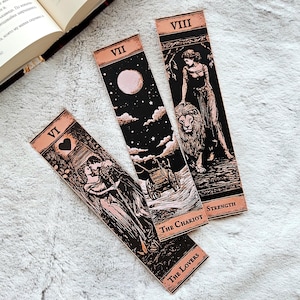 Puede incluir: Tres marcapáginas de tarot con ilustraciones en negro y rosa. Los marcapáginas presentan imágenes de The Lovers, The Chariot y Strength. Cada marcapáginas tiene un encabezado rosa con números romanos y el nombre de la carta.