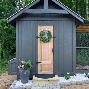 Puede incluir: Un gallinero de madera gris con una puerta de madera y una corona en la puerta. El gallinero tiene una peque&ntilde;a ventana y un techo de metal. Hay un camino de piedra que conduce al gallinero.
