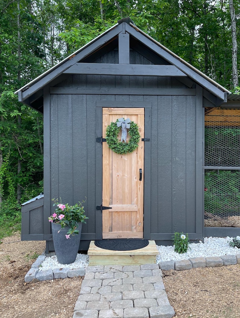 Puede incluir: Un gallinero de madera gris con una puerta de madera y una corona en la puerta. El gallinero tiene una peque&ntilde;a ventana y un techo de metal. Hay un camino de piedra que conduce al gallinero.